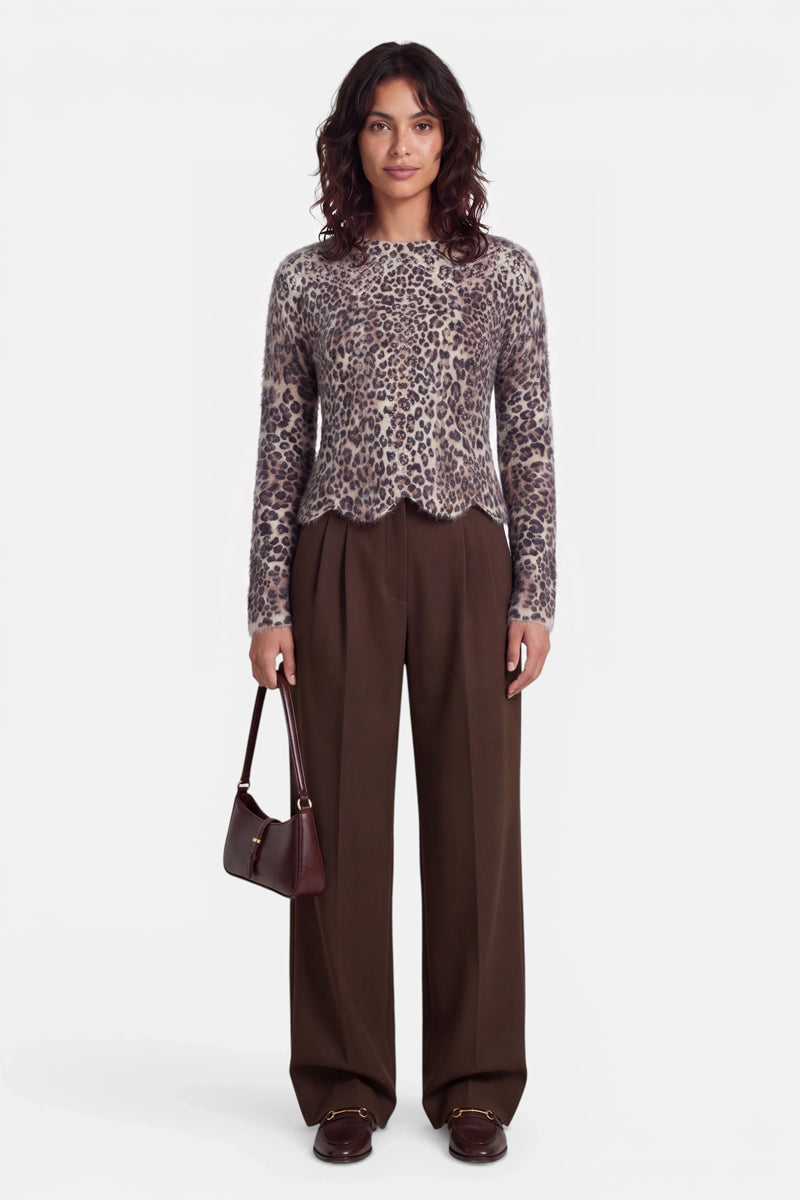 Eine Frau steht vor einem weißen Hintergrund und trägt einen langärmeligen Strickpullover mit Leopardenmuster und elegantem Wellenrand, eine braune Hose mit weitem Bein sowie braune Loafer. Sie hält eine kleine dunkelbraune Handtasche in der Hand und hat welliges, dunkles, schulterlanges Haar. Der Pullover besteht aus einer weichen Wolle-Alpaka-Mischung für zusätzlichen Tragekomfort. Sie blickt nach vorne und lächelt nicht.