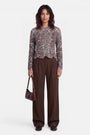 Eine Frau steht vor einem weißen Hintergrund und trägt einen langärmeligen Strickpullover mit Leopardenmuster und elegantem Wellenrand, eine braune Hose mit weitem Bein sowie braune Loafer. Sie hält eine kleine dunkelbraune Handtasche in der Hand und hat welliges, dunkles, schulterlanges Haar. Der Pullover besteht aus einer weichen Wolle-Alpaka-Mischung für zusätzlichen Tragekomfort. Sie blickt nach vorne und lächelt nicht.