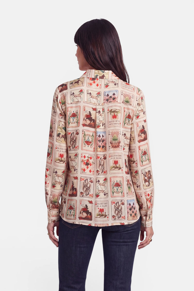 Eine Person mit langen, dunklen Haaren steht mit dem Rücken zur Kamera und trägt eine beige Hemdbluse mit bunten Vintage-Motiven aus Obst und Blumen. Ein Arm ist leicht angewinkelt, und die Person trägt eine dunkle Jeans. Der Hintergrund ist schlicht weiß.