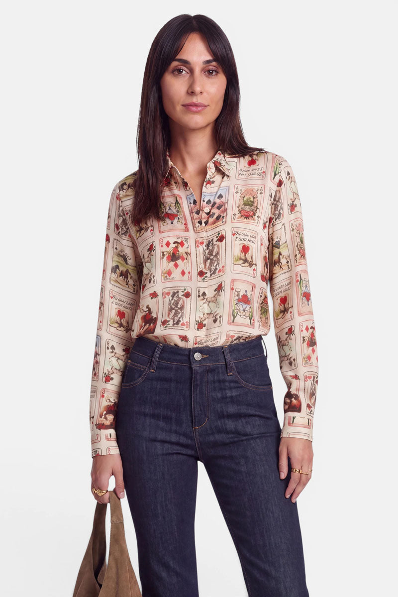 Eine Frau mit langen, dunklen Haaren trägt eine cremefarbene Hemdbluse, die mit bunten Blumen- und Pflanzenmotiven verziert ist, dazu eine dunkelblaue Jeans mit hoher Taille. In der linken Hand hält sie eine hellbraune Tasche und steht vor einem weißen Hintergrund.