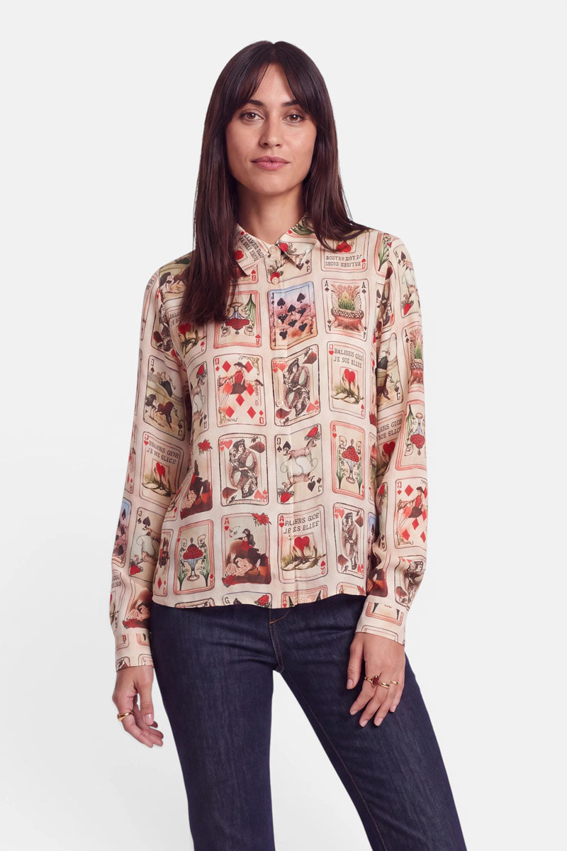 Eine Frau mit glattem braunem Haar trägt eine Hemdbluse mit einem Vintage-Spielkartenmuster, das verschiedene Illustrationen und Zahlen zeigt. Sie kombiniert die Bluse mit dem „Western-Karten“-Print mit einer dunklen Jeans, steht selbstbewusst da und blickt mit neutralem Gesichtsausdruck vor einem schlichten weißen Hintergrund in die Kamera.