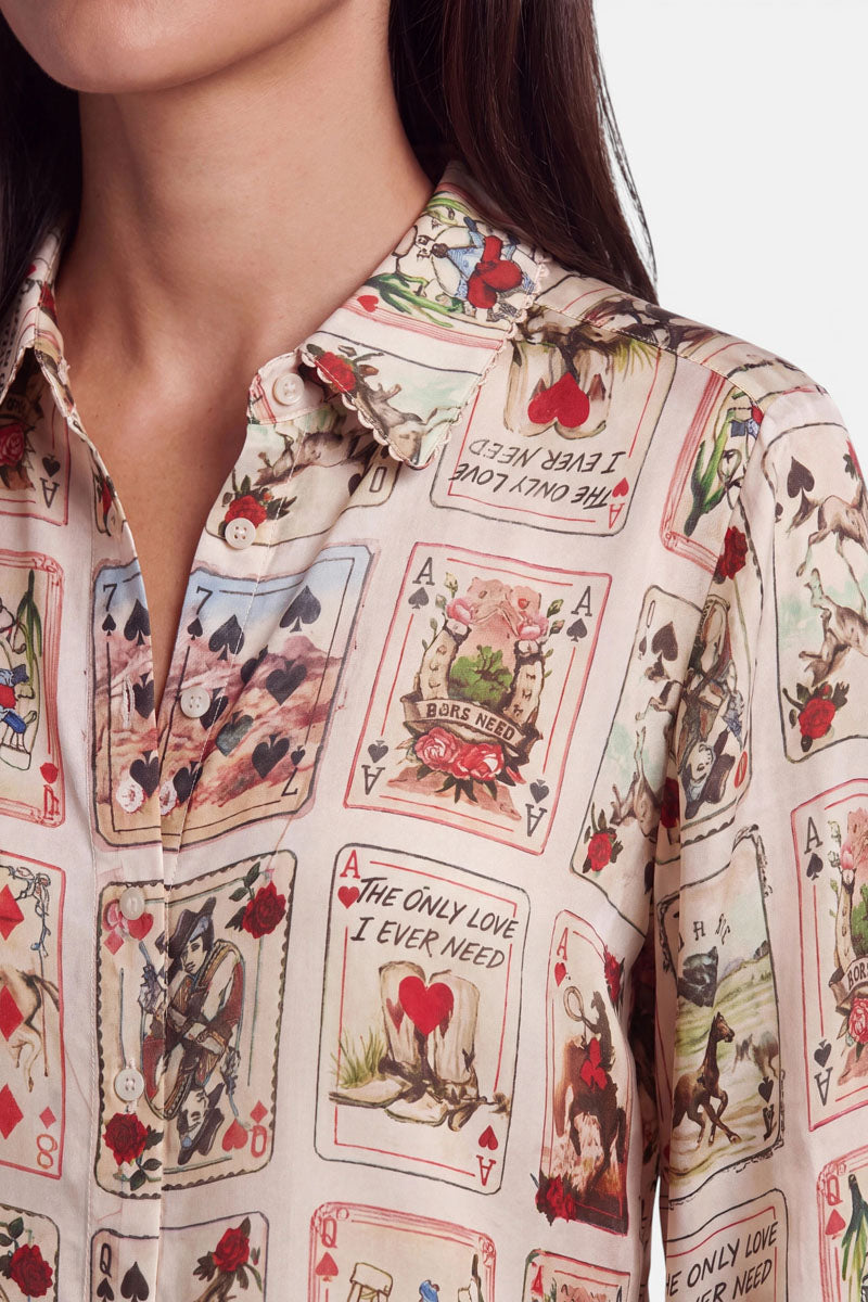 Eine Frau trägt eine cremefarbene Hemdbluse, die mit Vintage-Spielkarten, Rosen und romantischen Motiven verziert ist. Auf einigen Karten sind Herzen und Illustrationen zum Thema Liebe zu sehen, beispielsweise Paare und Sprüche wie „The only love I ever need“. Der Western-Karten-Print hebt sich wunderschön vom schlichten weißen Hintergrund ab.