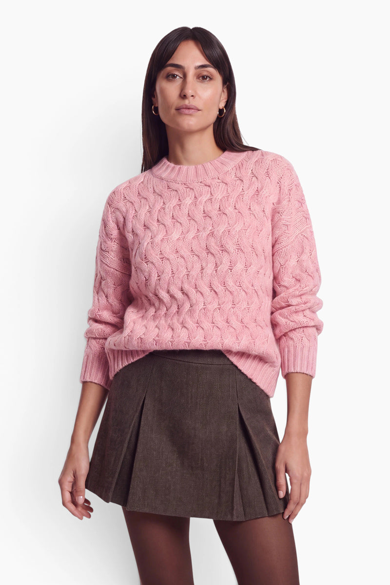 Eine Frau mit dunklem Haar trägt einen bubblegum-rosa, locker sitzenden Pullover mit Zopfmuster über einem dunkelbraunen, plissierten Minirock. Sie steht vor einem schlichten, cremefarbenen Studiohintergrund.