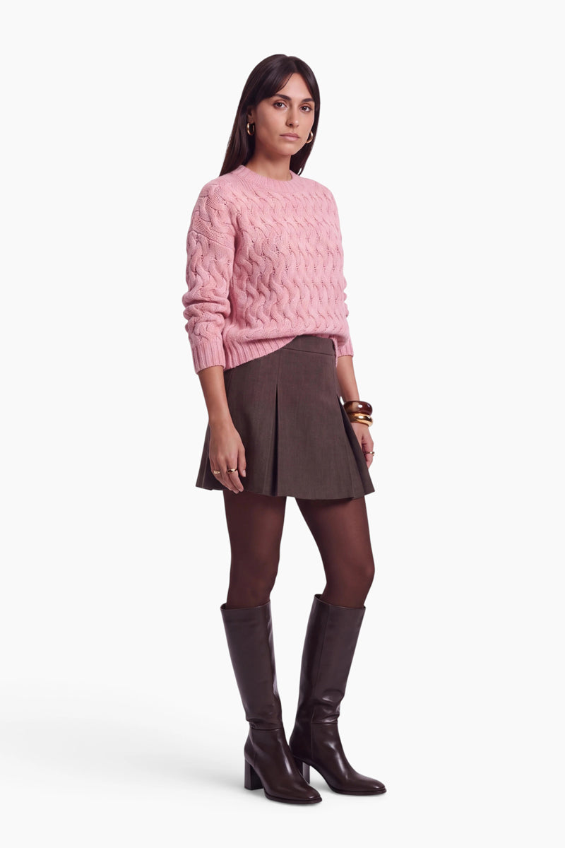 Vor einem weißen Hintergrund steht eine Frau mit dunklem Haar, goldenen Creolen und Ringen. Sie trägt einen rosa, locker sitzenden Pullover mit Zopfmuster, einen braunen Minirock mit Falten, dunkle Strumpfhosen und kniehohe Lederstiefel.