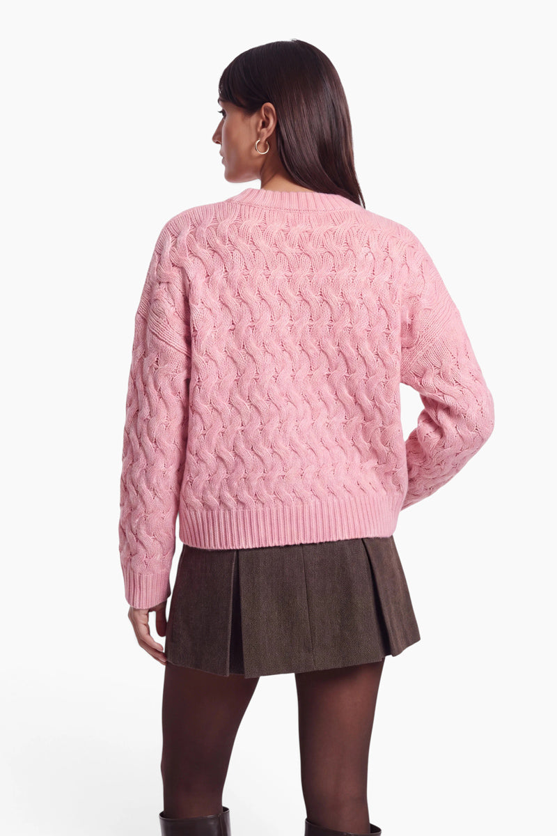 Vor einem schlichten weißen Hintergrund ist eine Frau von hinten zu sehen, die einen hellrosa, locker sitzenden Pullover mit Zopfmuster und Rundhalsausschnitt, einen dunkelbraunen, plissierten Minirock, dunkle Strumpfhosen und hohe Stiefel trägt.