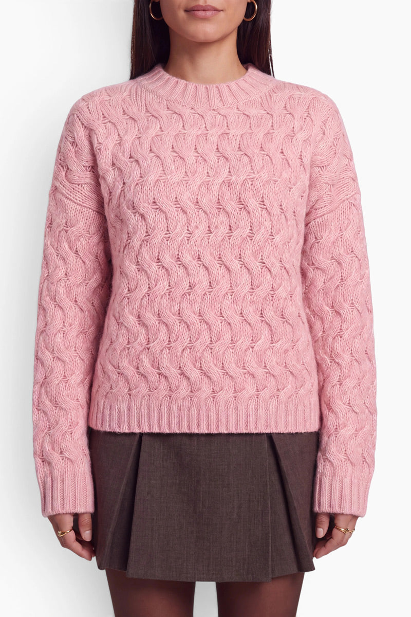 Eine Person, die einen weichen, bubblegum-rosa Zopfmusterpullover mit lockerer Passform und geripptem Rundhalsausschnitt trägt. Der Pullover wird mit einem dunkelbraunen, plissierten Minirock kombiniert.