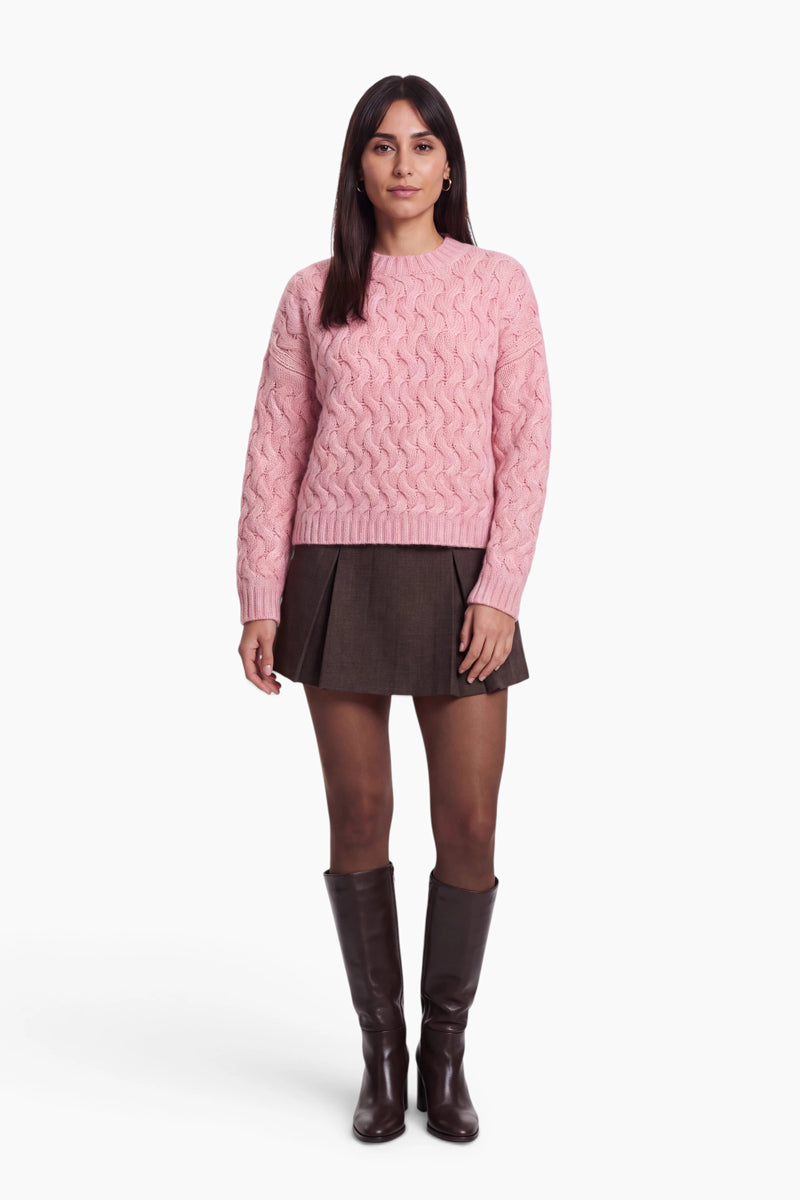 Eine Frau mit dunklem Haar steht mit dem Blick nach vorne und trägt einen rosa, locker sitzenden Zopfmusterpullover, einen dunkelbraunen Minirock mit Falten, eine transparente schwarze Strumpfhose und braune kniehohe Stiefel. Vor einem schlichten weißen Hintergrund hat sie einen neutralen Gesichtsausdruck.