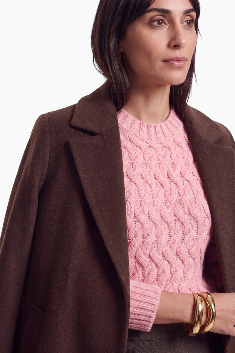 Eine Frau mit dunklem Haar trägt einen weichen braunen Wollmantel über einem locker sitzenden rosa Zopfmusterpullover. An ihren Handgelenken trägt sie goldene Armreifen und steht vor einem schlichten weißen Hintergrund.