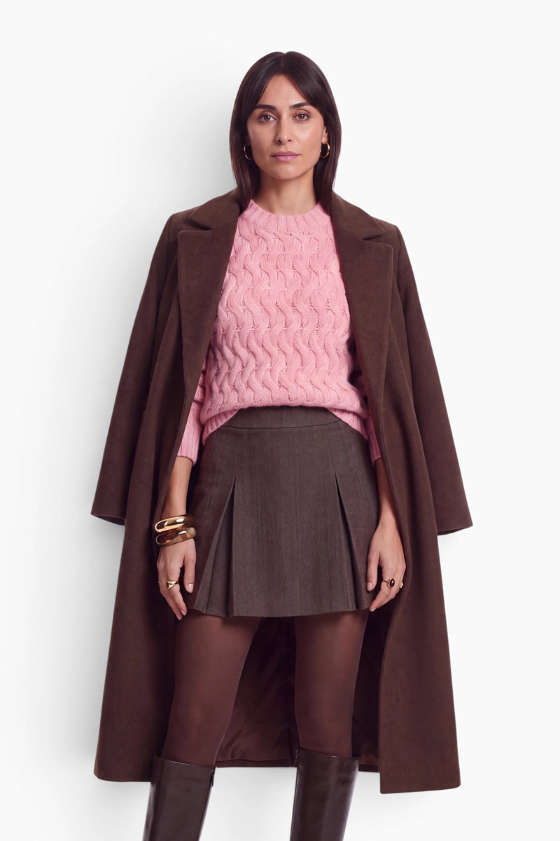 Vor einem weißen Hintergrund steht eine Frau mit dunklem Haar in einem locker sitzenden rosa Zopfmusterpullover, einem grauen Plissee-Minirock und einem braunen Mantel, den sie sich über die Schultern gelegt hat. Sie trägt braune Strumpfhosen und Stiefel sowie goldene Armreifen und Ringe als Accessoires.