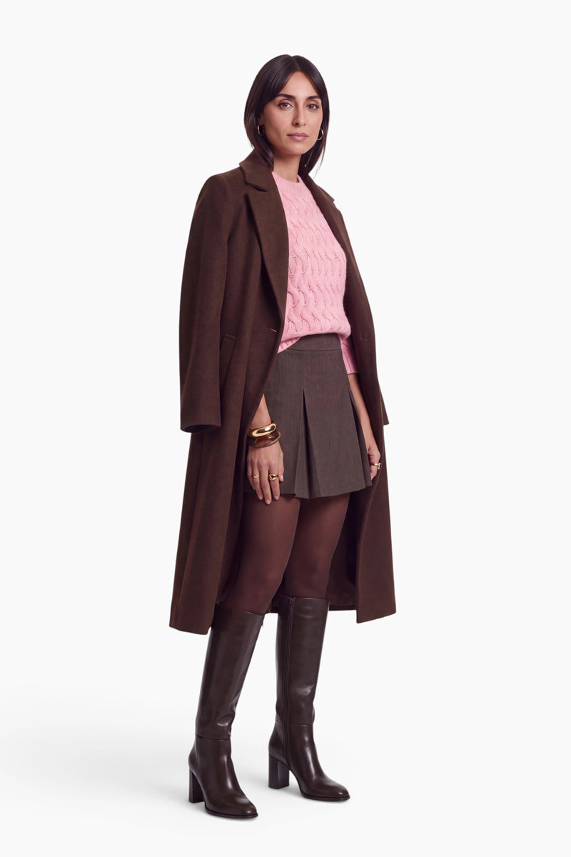 Eine Frau trägt einen langen braunen Mantel über einem rosa, locker sitzenden Zopfmusterpullover und einem dunklen, plissierten Minirock. Vor einem schlichten weißen Hintergrund rundet sie den Look mit braunen Strumpfhosen und kniehohen Lederstiefeln ab.
