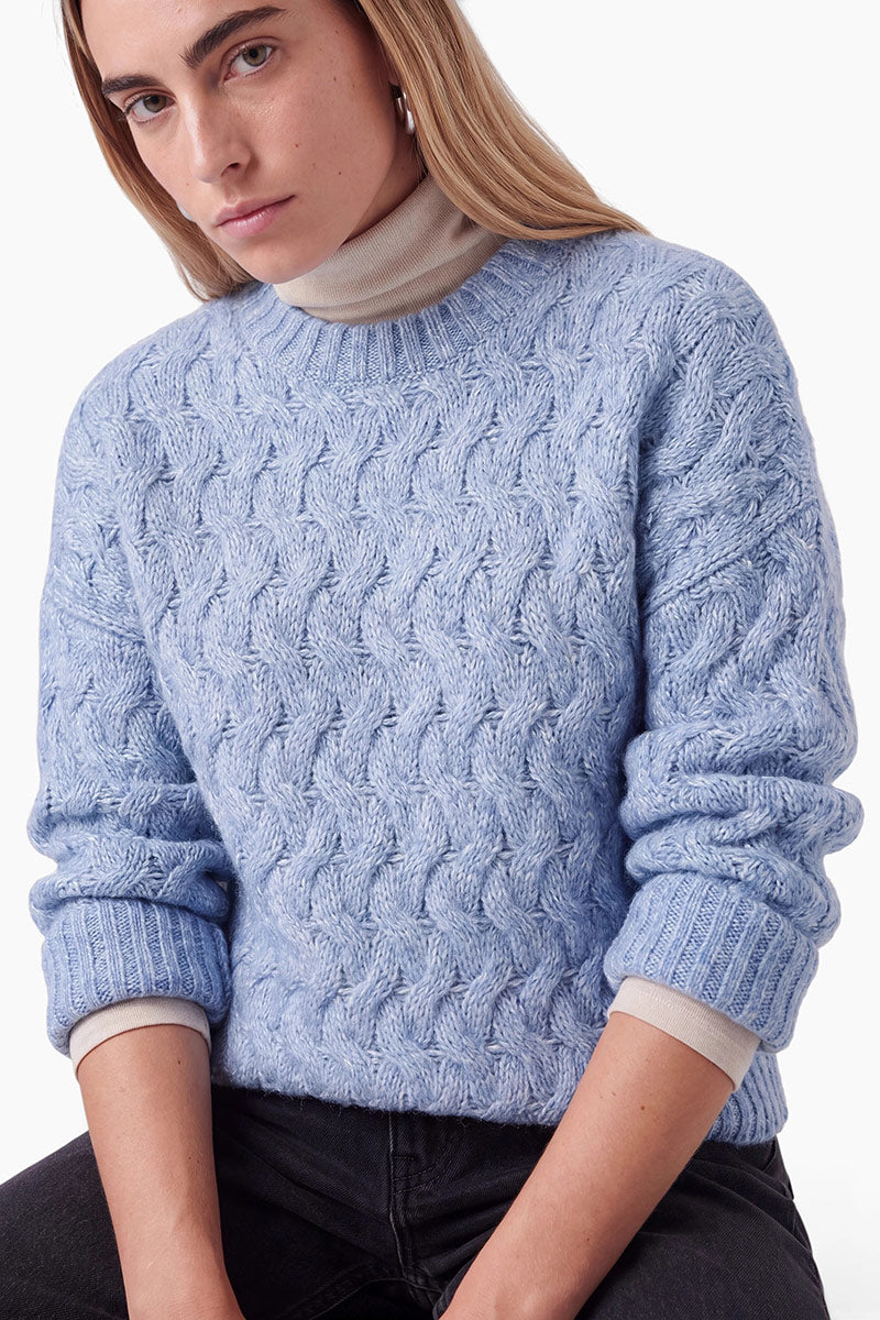Eine Person mit langen blonden Haaren trägt einen hellblauen, locker sitzenden Pullover mit Zopfmuster über einem beigen Rollkragenpullover. Sie hat einen neutralen Gesichtsausdruck und ist in dunkle Jeans gekleidet, vor einem schlichten weißen Hintergrund.