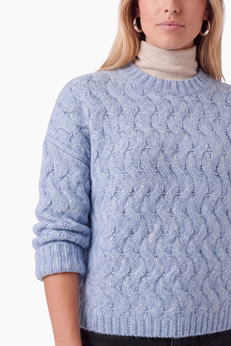 Eine blonde Person trägt einen hellblauen, strukturierten Zopfmusterpullover über einem beigen Rollkragenpullover. Der Pullover ist von der Brust aufwärts vor einem schlichten weißen Hintergrund abgebildet, wobei silberne Creolen zu sehen sind. Dieser grob gestrickte Pullover zeichnet sich durch eine bequeme, lockere Passform aus.