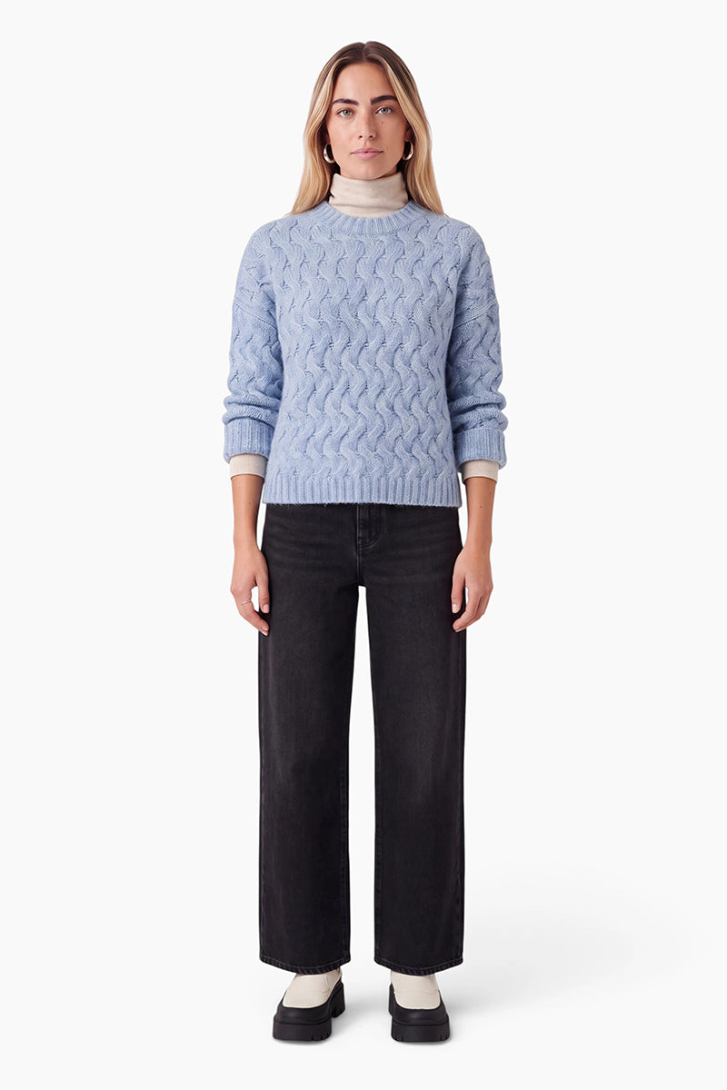 Eine Frau mit schulterlangen blonden Haaren trägt einen hellblauen Grober-Strickpullover über einem beigen Rollkragenpullover. Dazu kombiniert sie dunkelgraue Jeans mit weitem Bein und schwarze Plateau-Loafer und steht vor einem schlichten weißen Hintergrund.