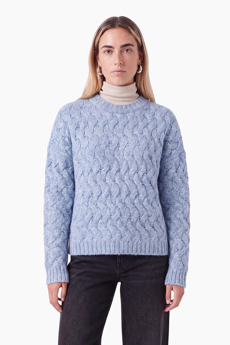 Eine blonde Frau, die einen hellblauen Zopfmusterpullover über einem beigen Rollkragenpullover trägt. Sie blickt vor einem schlichten weißen Hintergrund geradeaus und trägt dazu kleine silberne Creolen sowie eine schwarze Jeans.