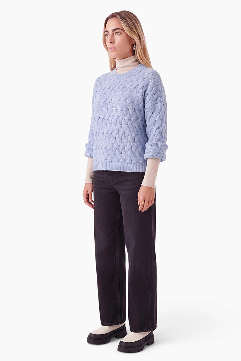 Eine Frau mit langen blonden Haaren trägt einen hellblauen Pullover mit Zopfmuster über einem beigen Rollkragenpullover, dazu eine schwarze Jeans mit weitem Bein. Sie steht vor einem schlichten weißen Hintergrund und trägt schwarz-weiße Loafer mit dicker Sohle.