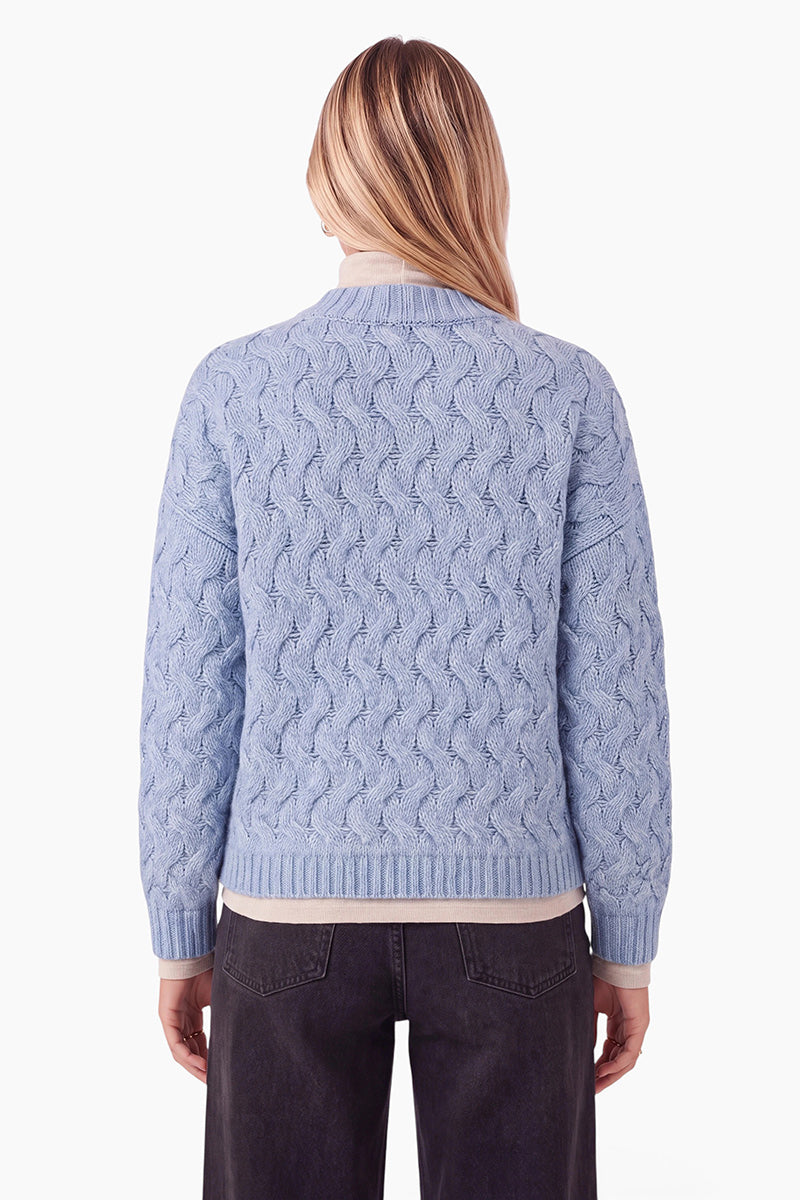 Eine Person mit blonden Haaren ist von hinten zu sehen. Sie trägt einen hellblauen, locker sitzenden Pullover mit Zopfmuster über einem cremefarbenen Hemd. Außerdem trägt sie dunkelgraue Jeans vor einem schlichten weißen Hintergrund.