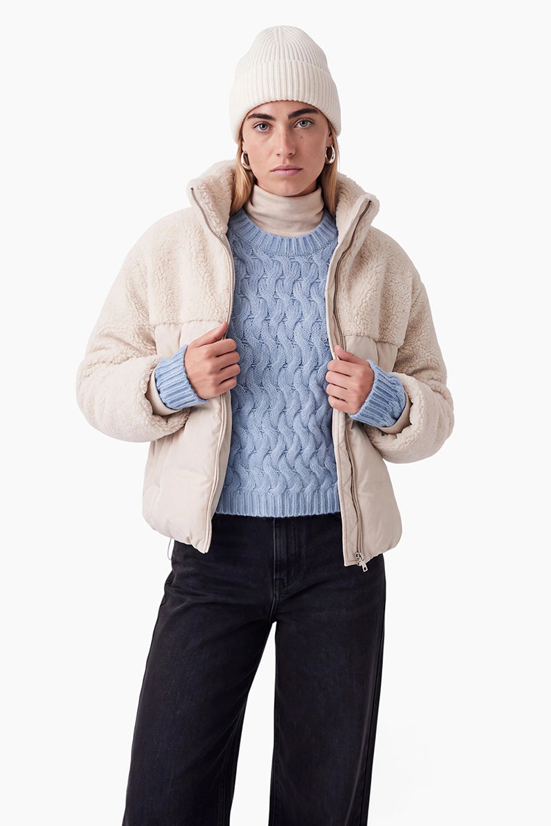 Eine Person trägt eine cremefarbene Mütze, einen hellblauen Pullover mit Zopfmuster und eine beige Daunenjacke mit Sherpa-Futter. Außerdem trägt sie eine dunkle Jeans und goldene Creolen vor einem schlichten weißen Hintergrund.
