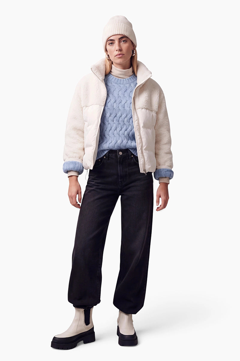 Eine Frau präsentiert eine cremefarbene Pufferjacke über einem hellblauen Pullover mit Zopfmuster. Vor einem weißen Hintergrund trägt sie eine dazu passende cremefarbene Mütze, schwarze Jeans mit weitem Bein und zweifarbige, creme-schwarze Chelsea-Stiefel.
