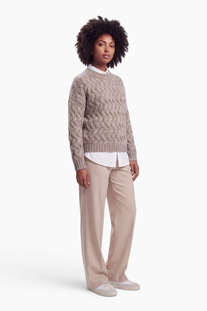 Eine Frau mit dunklem, voluminösem Lockenhaar trägt einen hellbraunen, grob gestrickten Zopfmusterpullover über einem weißen Hemd. Sie trägt eine hellbeige Hose und Sneaker in neutralen Farbtönen vor einem schlichten weißen Hintergrund.