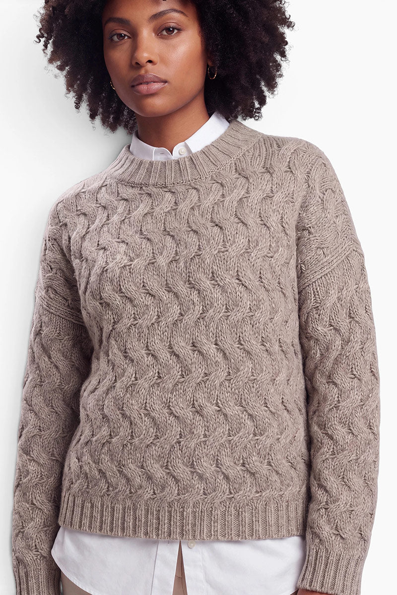 Eine Person mit dunklem, lockigem Haar trägt einen taupefarbenen, locker sitzenden Pullover mit Zopfmuster über einem weißen Hemd mit Kragen. Sie hat einen neutralen Gesichtsausdruck und blickt vor einem schlichten weißen Hintergrund direkt in die Kamera.