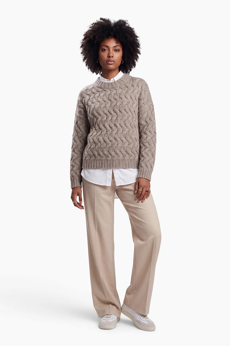 Eine Frau mit dunklem, lockigem Haar trägt einen beigen, locker sitzenden Pullover mit Zopfmuster über einem weißen Hemd mit Kragen. Sie steht vor einem schlichten weißen Hintergrund und trägt dazu eine beige Hose und weiße Turnschuhe.