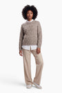 Eine Frau mit dunklem, lockigem Haar trägt einen beigen, locker sitzenden Pullover mit Zopfmuster über einem weißen Hemd mit Kragen. Sie steht vor einem schlichten weißen Hintergrund und trägt dazu eine beige Hose und weiße Turnschuhe.