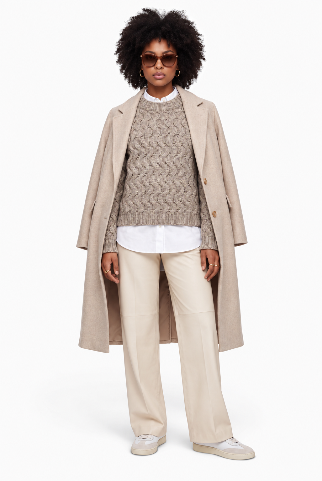 Vor einem schlichten weißen Hintergrund steht eine Frau mit dunklem Afro, die eine braune Sonnenbrille trägt, einen beigen, locker sitzenden Zopfstrickpullover über einem weißen Hemd, einen dazu passenden Mantel, der ihr über die Schultern drapiert ist, sowie eine cremefarbene Hose und helle Turnschuhe.