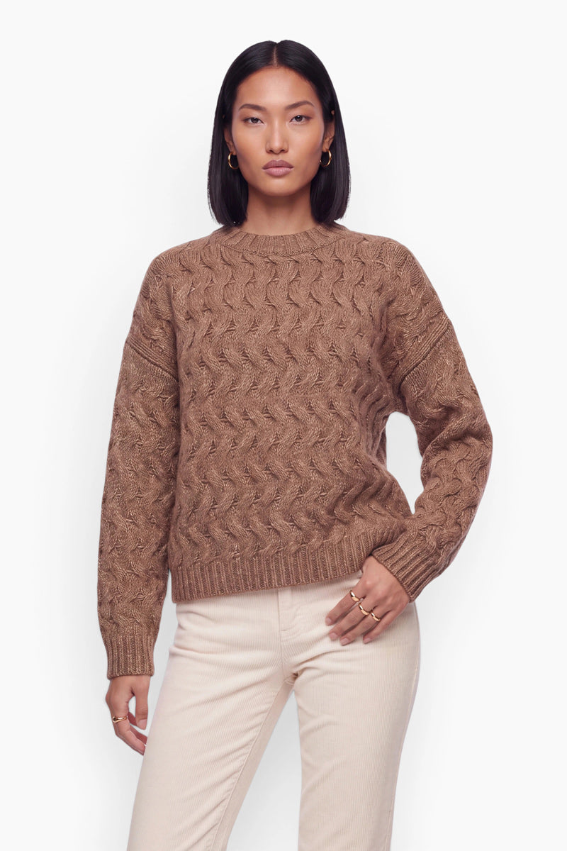 Eine Person mit dunklem Haar und goldenen Creolen trägt einen braunen, locker sitzenden Pullover mit Zopfmuster und eine cremefarbene Hose. Sie steht vor einem schlichten weißen Hintergrund und trägt an der linken Hand zwei goldene Ringe.