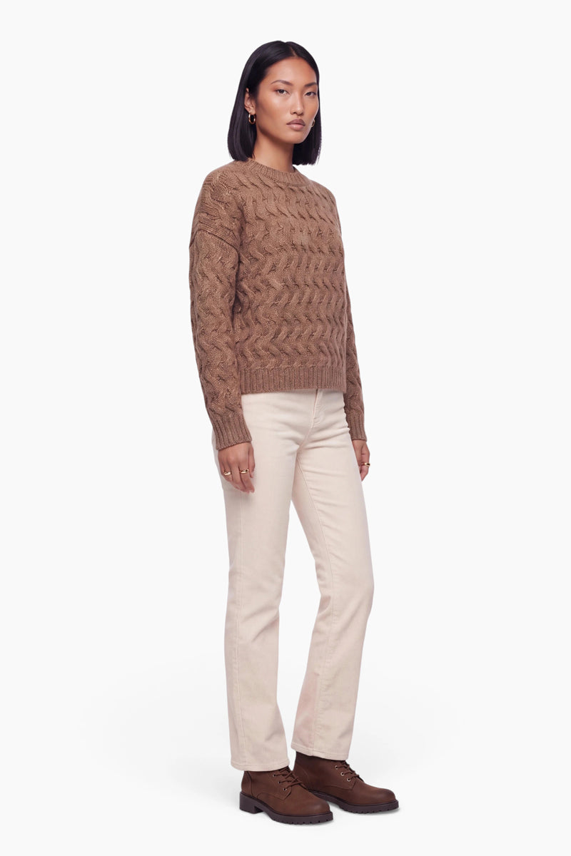 Eine Frau mit schulterlangem dunklem Haar trägt einen braunen, grob gestrickten Zopfmusterpullover, eine cremefarbene Hose mit geradem Bein und braune Schnürstiefel. Sie steht vor einem schlichten weißen Hintergrund und blickt in die Kamera.