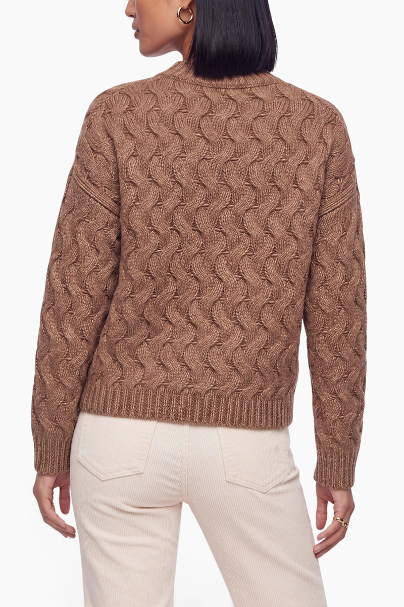 Eine von hinten gezeigte Person trägt einen braunen, langärmeligen Pullover mit Zopfmuster, der am Rundhalsausschnitt und am Saum gerippt ist. Dazu trägt sie eine helle Hose vor einem schlichten weißen Hintergrund.