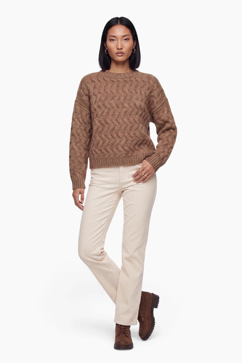 Eine Frau mit schulterlangem dunklem Haar präsentiert einen braunen, locker sitzenden Pullover mit Zopfmuster, eine hellcremefarbene Hose und braune Schnürstiefel. Sie steht vor einem schlichten weißen Hintergrund und hat die Hand in die Tasche gesteckt.