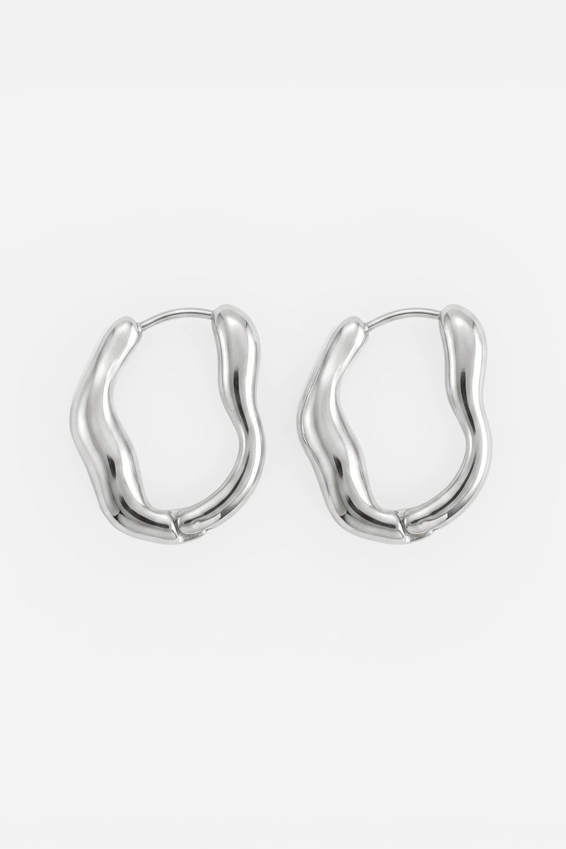 Ein Paar skulpturale Ohrringe von Princess Goes Hollywood aus Silber mit einer wellenförmigen, organischen Silhouette. Die aus Edelstahl gefertigten Creolen besticht durch ihre glatte, polierte Metalloberfläche, die einen markanten Akzent setzt. Jeder Ohrring ist oben mit einem Scharnierverschluss gesichert. Vor einem schlichten weißen Hintergrund kommen diese modernen Ohrringe mit ihrem unverwechselbaren und kunstvollen Design besonders gut zur Geltung.