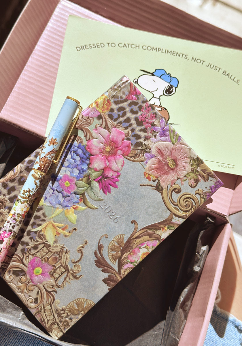 Ein stilvolles Set von Princess Goes Hollywood aus floral gemustertem Agenda und Kugelschreiber ruht auf rosa Seidenpapier in einer Box. Dahinter steht eine Snoopy-Karte mit „Dressed to catch compliments, not just balls“.