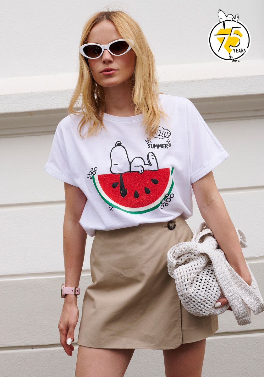 A blonde woman wears white cat-eye sunglasses, a Princess Goes Hollywood Snoopy-Print cotton T-shirt with Hello Summer text and Snoopy on a watermelon, beige high-waisted shorts, pink watch, chunky knit bag. 75 Years Snoopy logo top right.