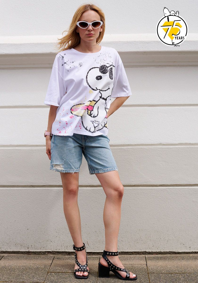 A woman in white cat-eye sunglasses wears a Princess Goes Hollywood weißes Baumwolle T-Shirt mit Snoopy-Print zum 75. Jubiläum, light denim knee-length shorts, and black sandals with Strassverzierung, posing by a wall with a yellow 75 Years Snoopy logo.
