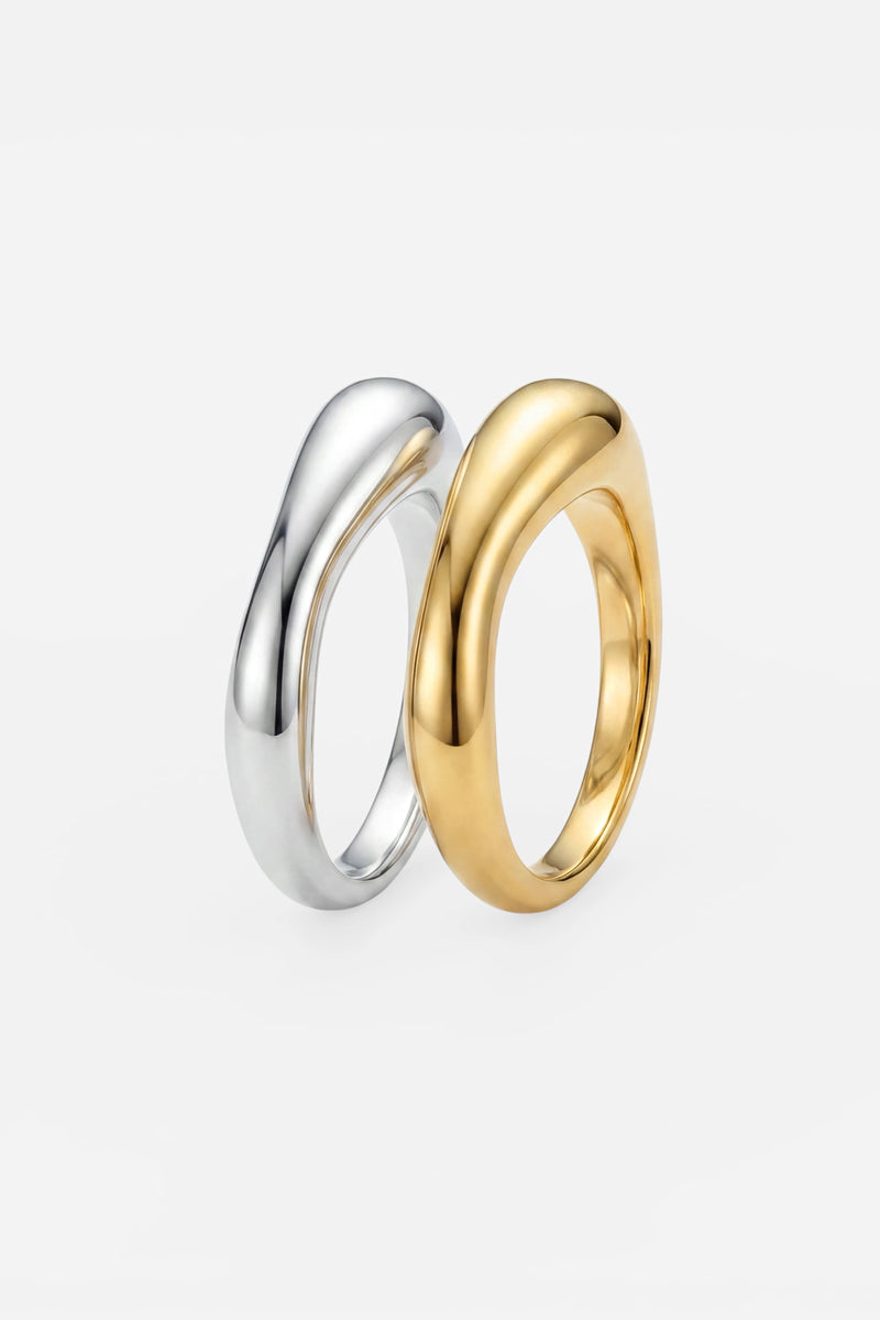 Zwei glatte, geschwungene Ringe von Princess Goes Hollywood stehen aufrecht auf einer weißen Unterlage. Der eine Ring ist aus 925er Sterlingsilber fein gearbeitet und mit einer glänzenden Silberoberfläche versehen, während der andere eine elegante 18-Karat-Vergoldung in glänzendem Gold aufweist. Beide zeichnen sich durch eine moderne, organische Form mit fließenden, asymmetrischen Linien aus, die einen zeitgemäßen und raffinierten Look schaffen.