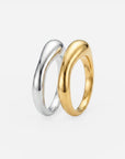 Zwei glatte, geschwungene Ringe von Princess Goes Hollywood stehen aufrecht auf einer weißen Unterlage. Der eine Ring ist aus 925er Sterlingsilber fein gearbeitet und mit einer glänzenden Silberoberfläche versehen, während der andere eine elegante 18-Karat-Vergoldung in glänzendem Gold aufweist. Beide zeichnen sich durch eine moderne, organische Form mit fließenden, asymmetrischen Linien aus, die einen zeitgemäßen und raffinierten Look schaffen.