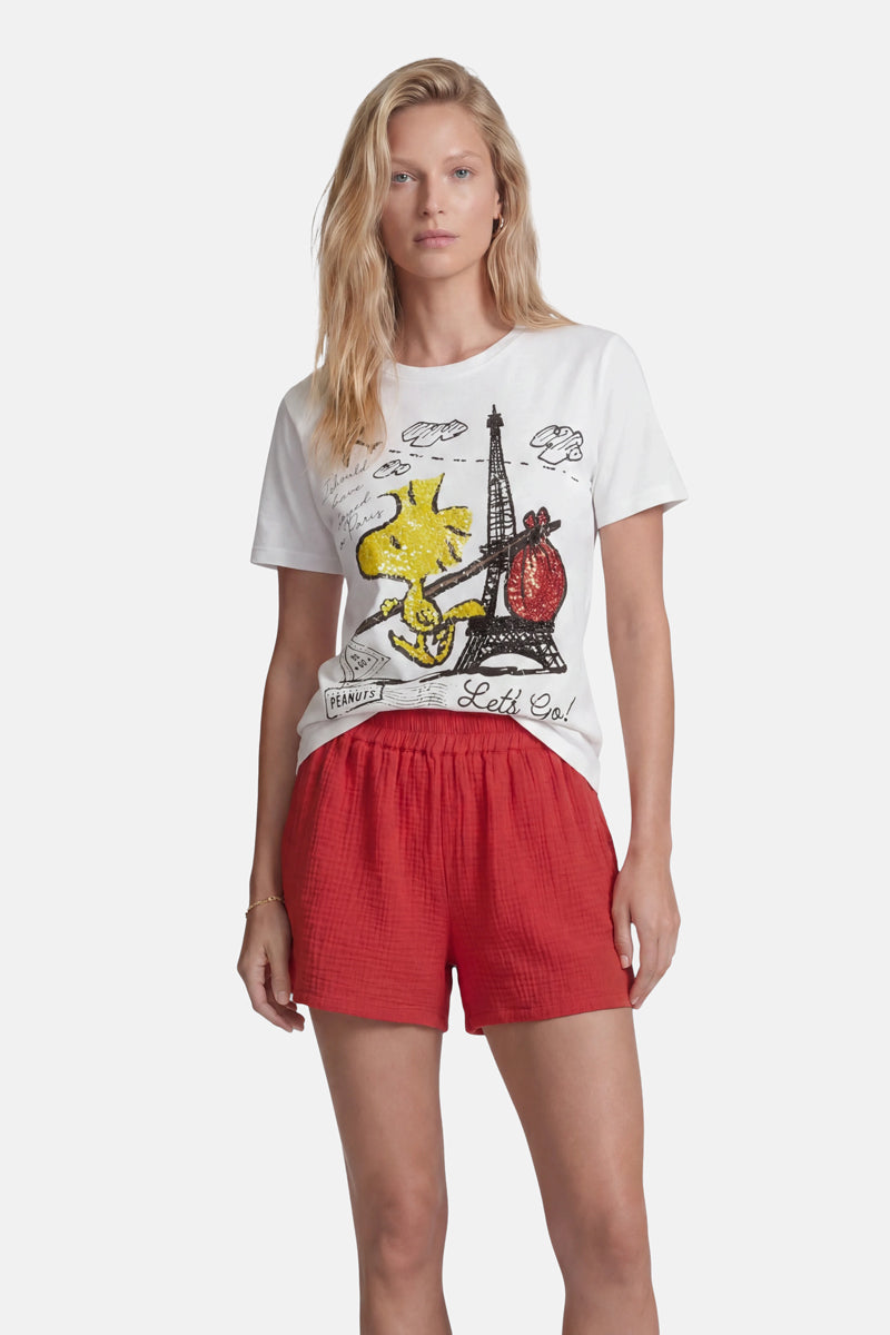 Eine Frau mit langen blonden Haaren präsentiert ein T-Shirt der Limited Edition „Princess Goes Hollywood“ mit Woodstock-Motiv, auf dem ein gelber Comic-Vogel, der Eiffelturm und der Schriftzug „Let’s Go!“ zu sehen sind, kombiniert mit weiten roten Shorts. Sie blickt vorwärts vor einem weißen Hintergrund.