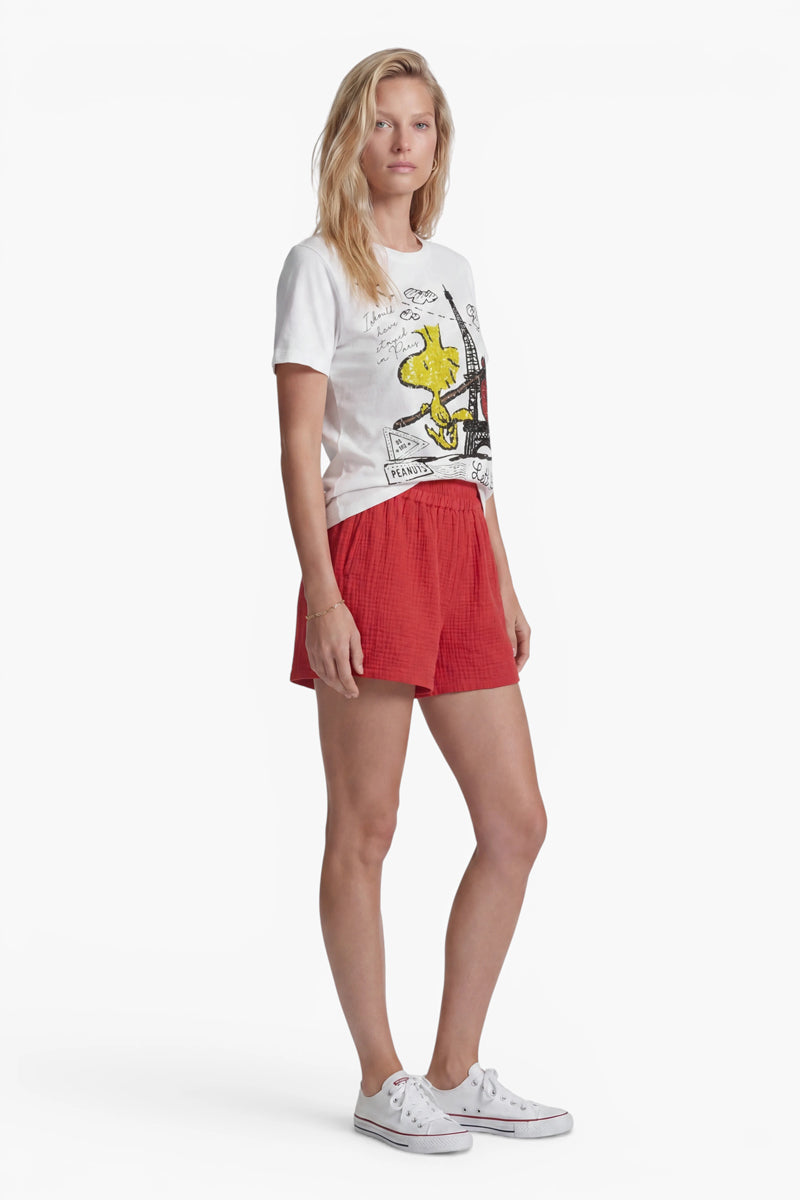 Vor einem weißen Hintergrund steht eine Frau mit heller Haut und langen blonden Haaren. Sie trägt ein T-Shirt der Limited Edition „Princess Goes Hollywood“ mit Woodstock-Aufdruck, auf dem eine gelbe Zeichentrickfigur abgebildet ist, dazu rote Shorts und weiße Turnschuhe. Ihr Gesichtsausdruck ist neutral, die Hände hält sie an den Seiten.