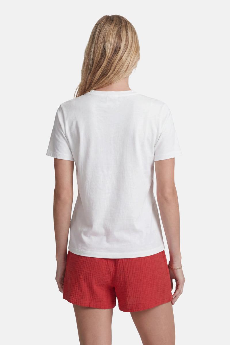 Eine Frau mit langen blonden Haaren steht mit dem Rücken zur Kamera und trägt ein schlichtes weißes T-Shirt der Limited Edition „Princess Goes Hollywood“ sowie rote Shorts mit Strukturmuster. Der Hintergrund ist weiß, und ihre Arme liegen entspannt an den Seiten.