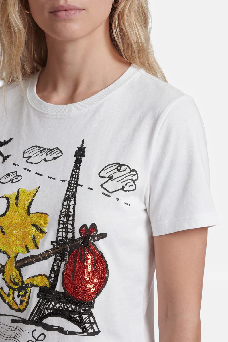 Eine Frau in einem weißen T-Shirt der Limited Edition „Princess Goes Hollywood“, auf dem eine gelbe Comic-Katze abgebildet ist, die eine rote, mit Pailletten besetzte Tasche hält, steht vor einer Skizze des Eiffelturms mit schwarzen Wolken. Nur ihr Kopf und ihr Oberkörper sind zu sehen.