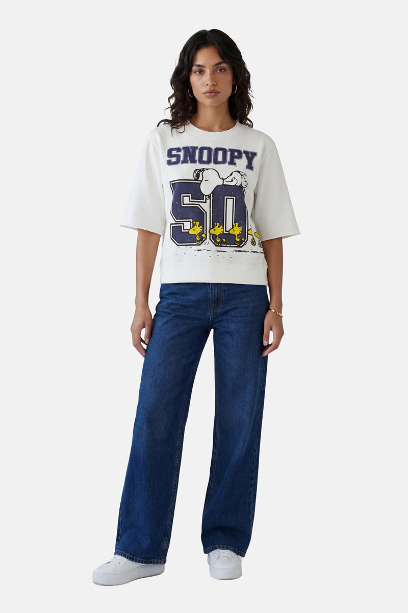 Eine Frau mit dunklem, gewelltem Haar und ausdruckslosem Gesicht trägt den Snoopy-Sweater aus Baumwolle aus der limitierten Auflage „Princess Goes Hollywood“, eine blaue Jeans mit weitem Bein und weiße Sneaker und steht vor einem schlichten, hellen Hintergrund.