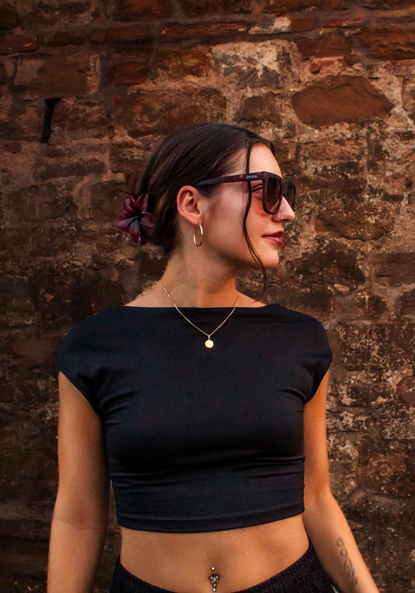 Eine Frau mit dunklem Haar in einem Dutt, geschmückt mit einer roten Blume, steht vor einer rustikalen Steinmauer. Sie trägt eine Okkia Oversized Sonnenbrille mit UV 400-Schutz, ein schwarzes Crop-Top, goldene Ohrringe, mehrlagige Ketten und ein Bauchnabelpiercing.
