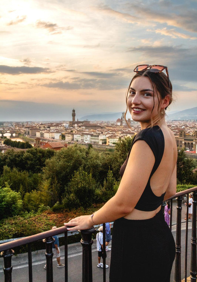 Eine Frau mit braunem Haar in einem tiefen Dutt, die ein schwarzes rückenfreies Kleid und eine übergroße Okkia-Sonnenbrille auf dem Kopf trägt, lächelt in die Kamera, während sie an einem Geländer mit Blick auf eine Stadtlandschaft im Sonnenuntergang steht.