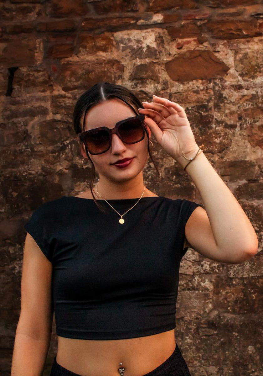 Eine Frau mit dunklem Haar in einem tiefen Dutt trägt eine Okkia Oversized Sonnenbrille mit UV 400-Schutz, ein schwarzes Crop-Top, goldene Halsketten und ein Armband. Sie steht selbstbewusst vor einer rustikalen Steinmauer, hält ihre Sonnenbrille und schaut in die Kamera.