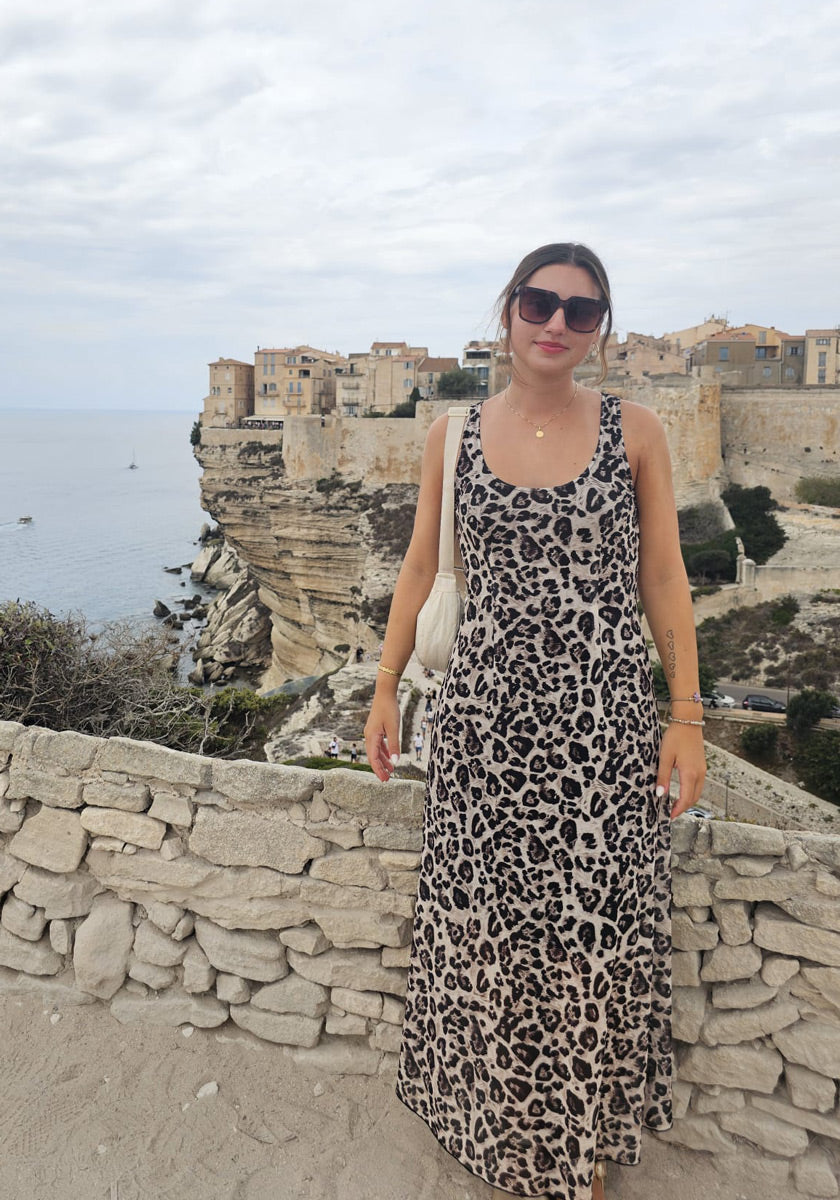 Eine Frau in einem ärmellosen Kleid mit Leopardenmuster und einer übergroßen Okkia-Sonnenbrille steht an einer Steinmauer mit Küstenklippen, historischen Gebäuden und dem Meer im Hintergrund. Sie trägt eine weiße Tasche auf der Schulter und lächelt in den wolkenverhangenen Himmel.