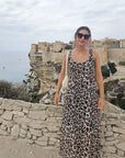 Eine Frau in einem ärmellosen Kleid mit Leopardenmuster und einer übergroßen Okkia-Sonnenbrille steht an einer Steinmauer mit Küstenklippen, historischen Gebäuden und dem Meer im Hintergrund. Sie trägt eine weiße Tasche auf der Schulter und lächelt in den wolkenverhangenen Himmel.