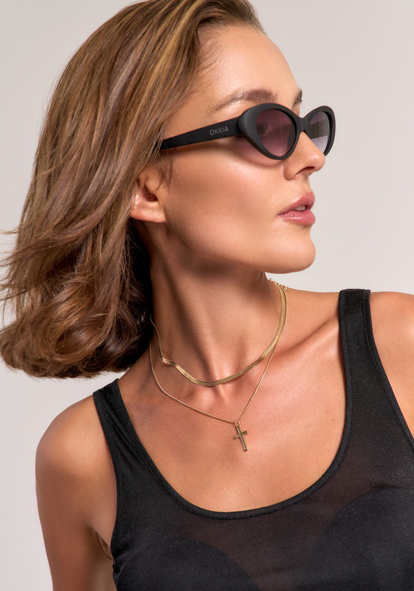 A woman with chin-length brown hair wears black Princess Goes Hollywood sunglasses, a black sleeveless top, and layered gold necklaces—a Schlangenkette and one with a Kreuzanhänger—looking to her right against a light background.
