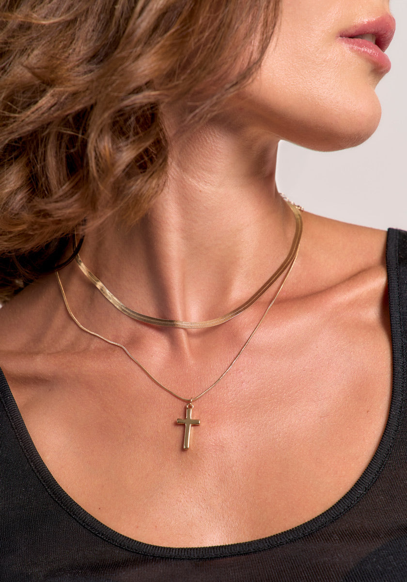 A woman with wavy brown hair wears a black top and two gold Princess Goes Hollywood necklaces—a Schlangenkette choker and a longer chain with a small Kreuzanhänger—against a neutral background, showing only her lower face and neck.