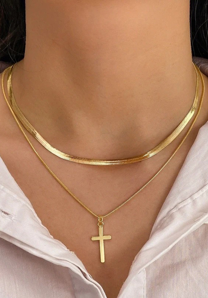 A close-up of a person’s neck wearing two gold Princess Goes Hollywood necklaces—a Schlangenkette choker and a thin Kette mit Kreuzanhänger—paired with a white collared shirt.